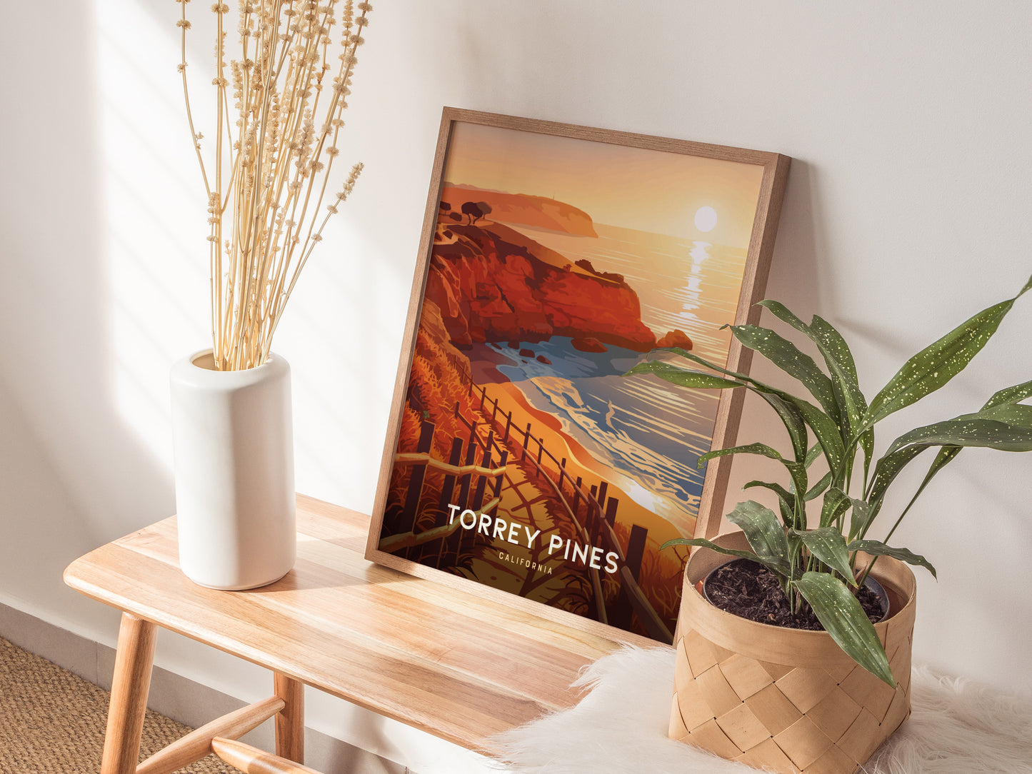 A framed Torrey Pines poster sits on a wooden bench, surrounded by a white vase with dried stems and a potted green plant, in a sunlit room.