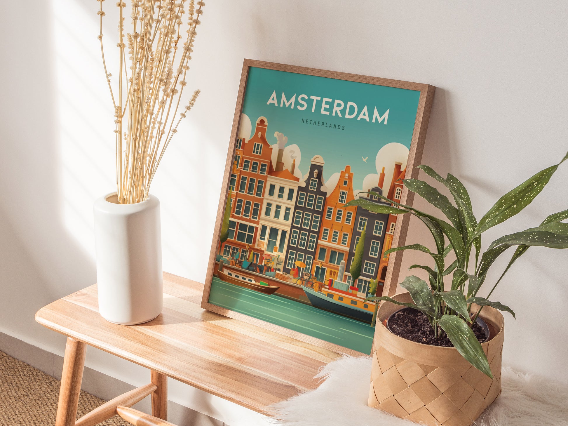 A framed Amsterdam poster with colorful canal houses is displayed on a wooden bench, alongside a white vase with dried flowers and a potted plant.