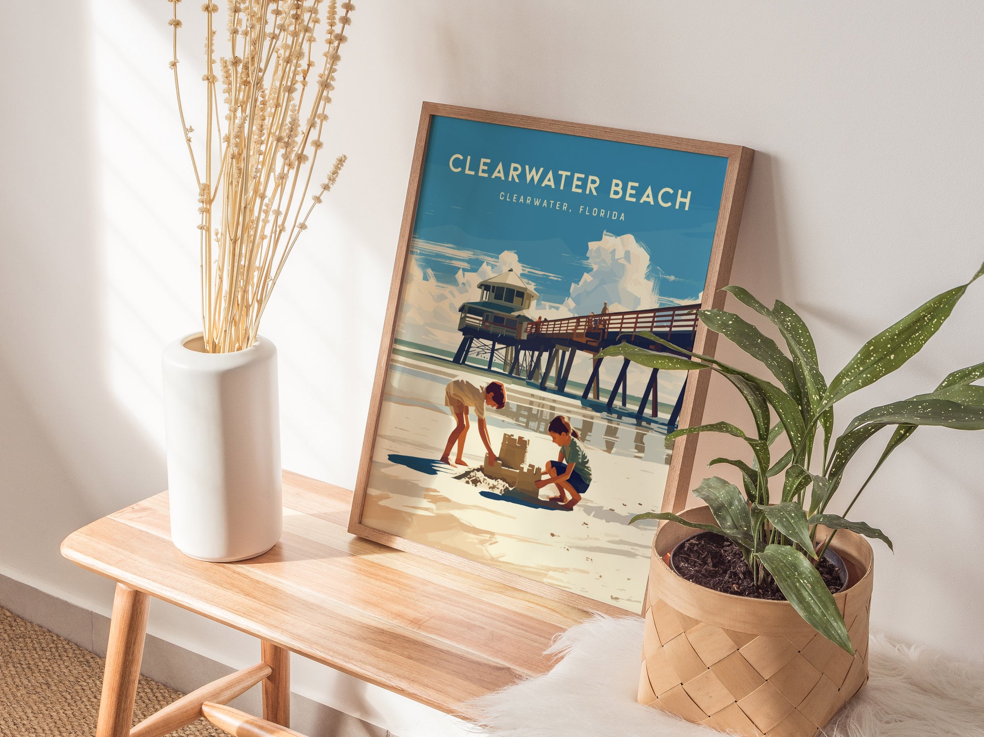 A framed Clearwater Beach poster rests on a wooden table, depicting children building a sandcastle under a lifeguard tower. Nearby are a white vase with dried plants and a leafy.