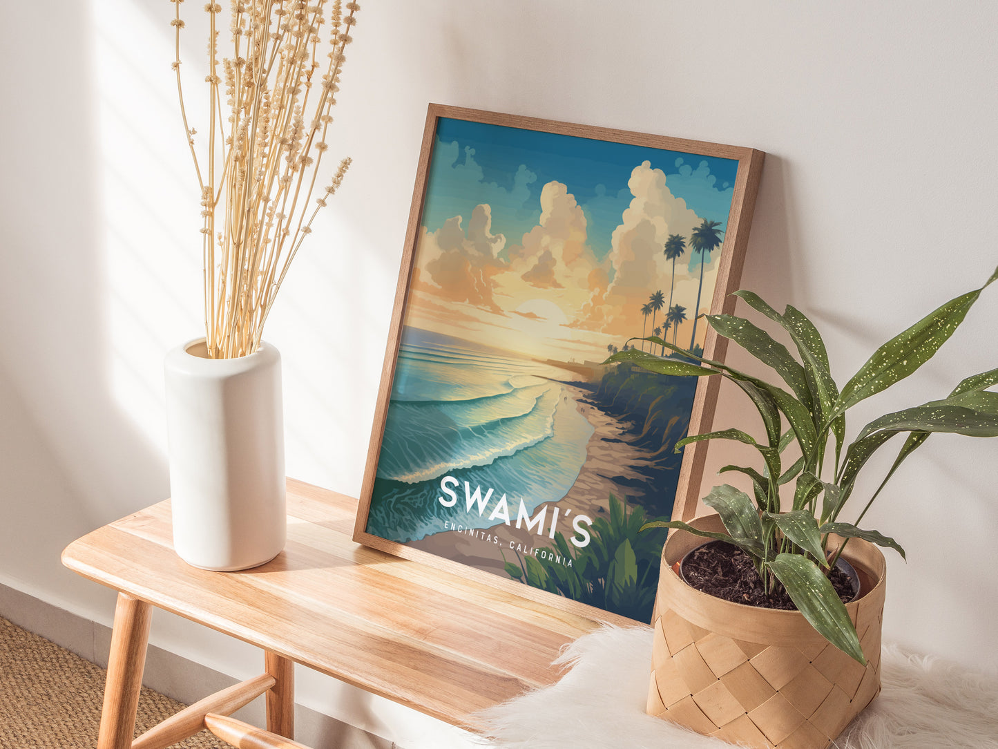 A framed poster of Swamis Encinitas surf spot with a sunset view rests on a wooden bench, accompanied by a white vase with dried flowers and a potted plant.