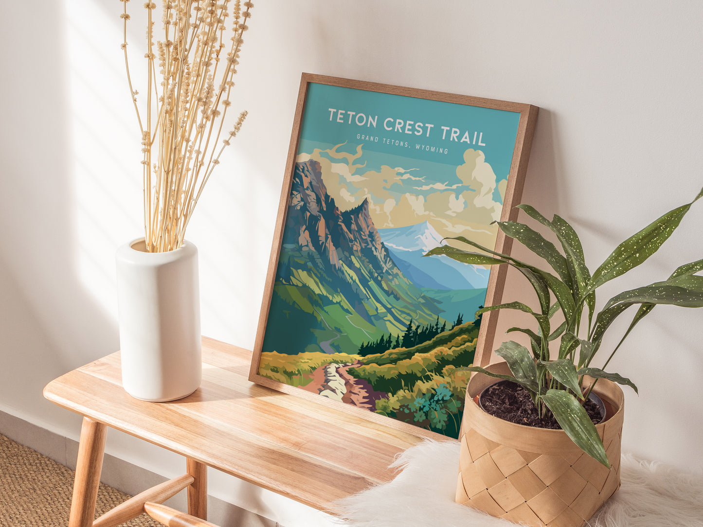 A framed Teton Crest Trail poster rests on a wooden bench, surrounded by a white vase with dried plants and a potted green plant. Natural light illuminates the scene.