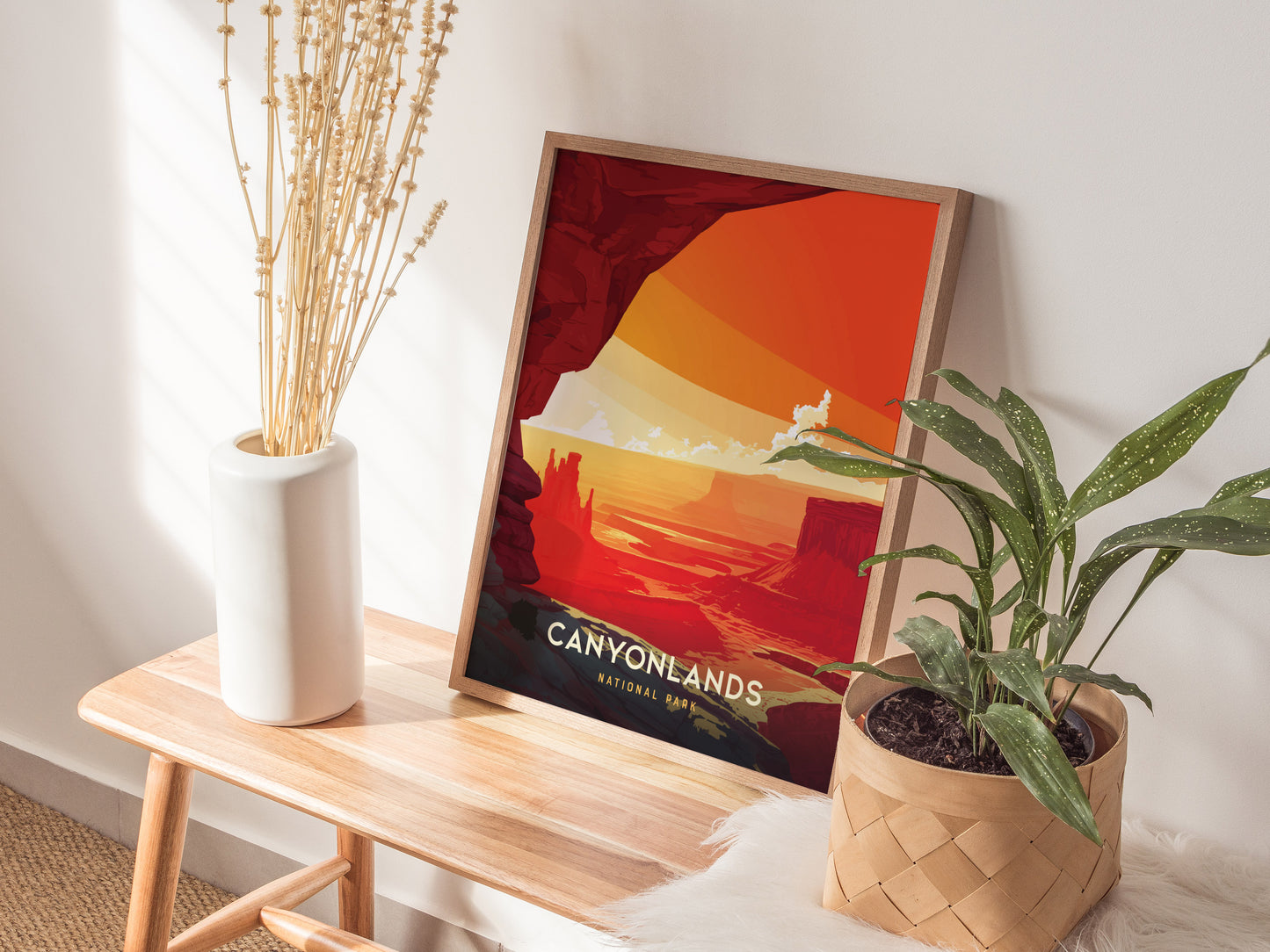 A framed Canyonlands National Park poster with vibrant orange and red hues rests on a wooden bench between a white vase with dried plants and a green potted plant.