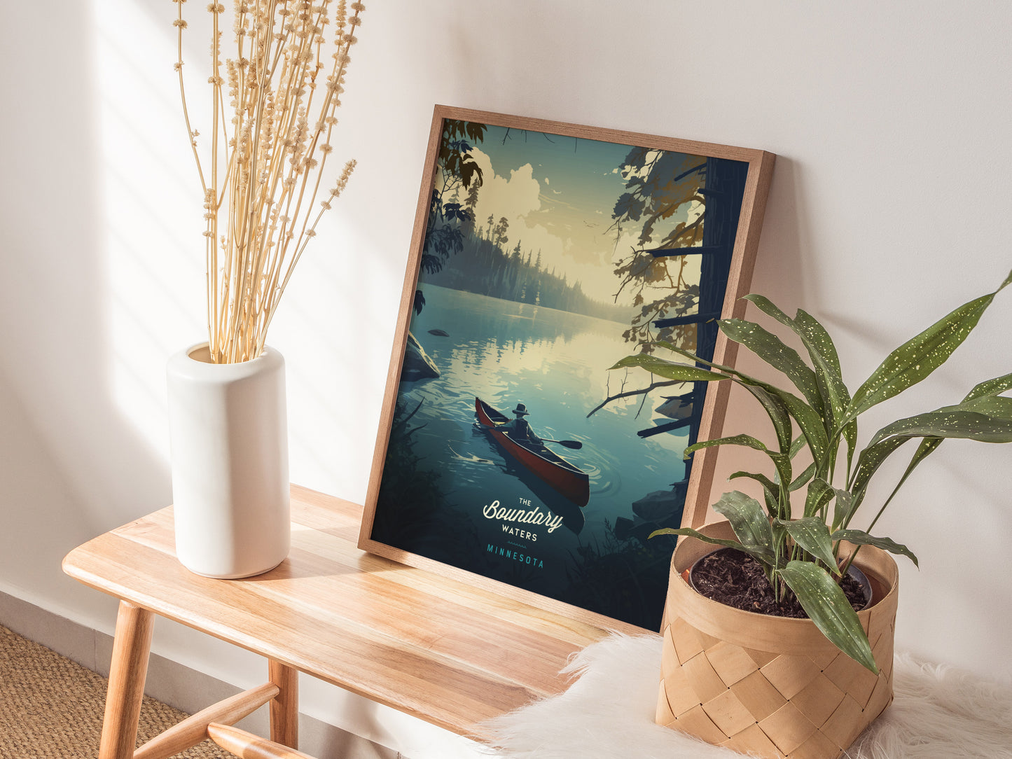 A framed poster of a canoe scene rests on a wooden table, accompanied by a white vase with dried flowers and a potted plant in a woven basket.