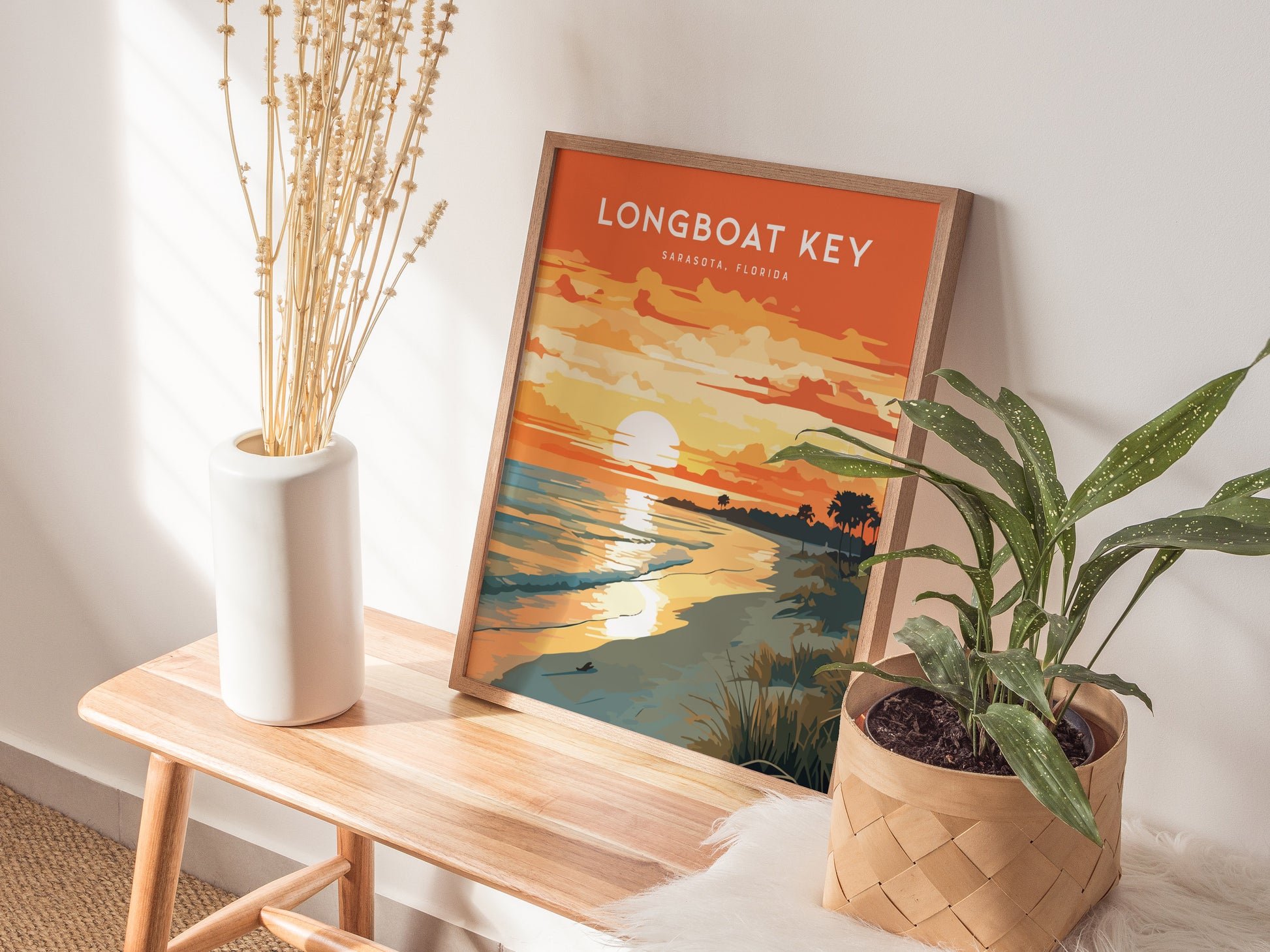A framed Longboat Key poster featuring a vibrant sunset over the ocean is displayed on a wooden bench. Next to it, a white vase with dried stems.