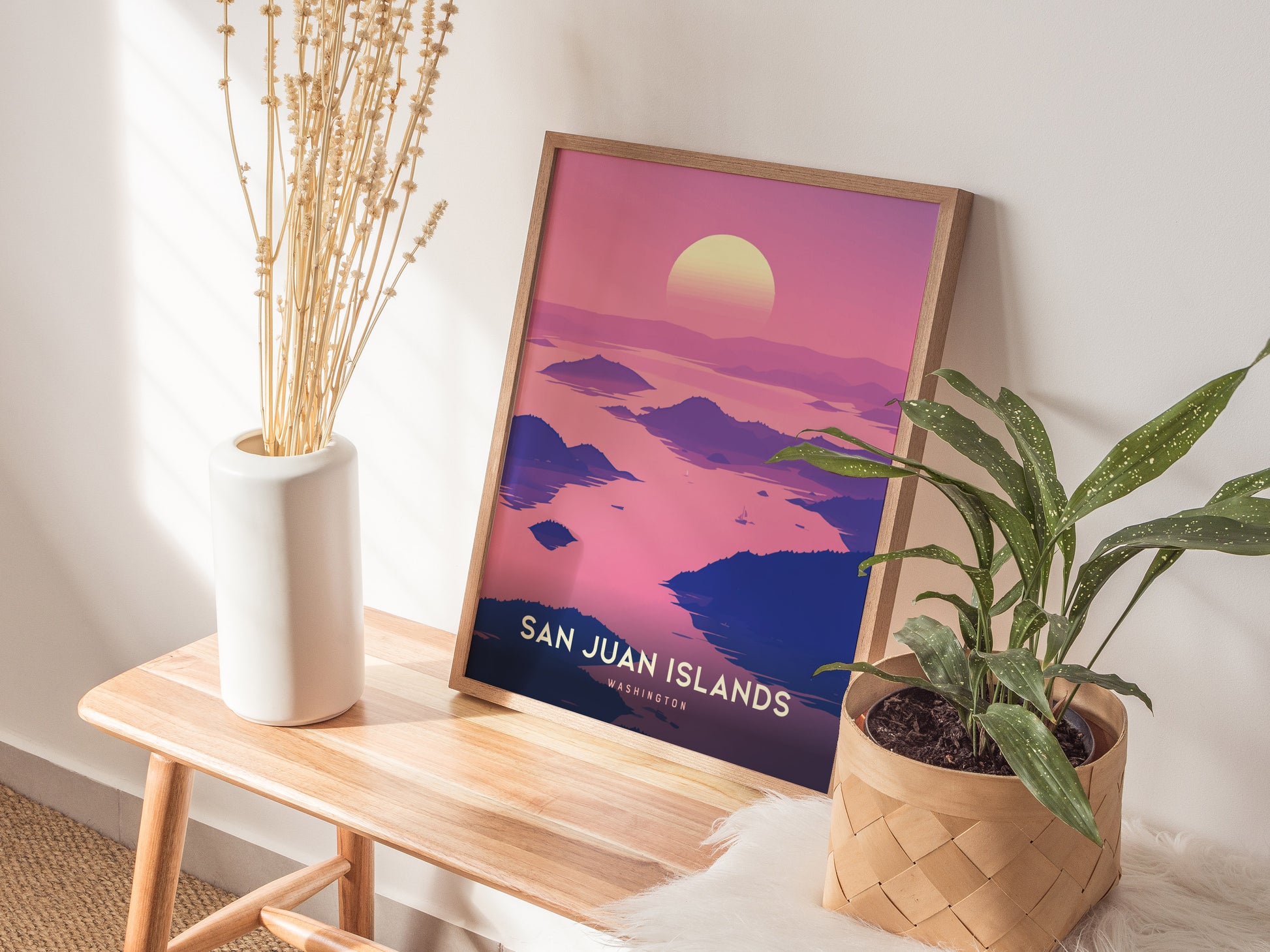 A framed San Juan Islands poster with a vibrant pink and purple sunset is displayed on a wooden table, accompanied by a white vase with dried flowers and a potted.