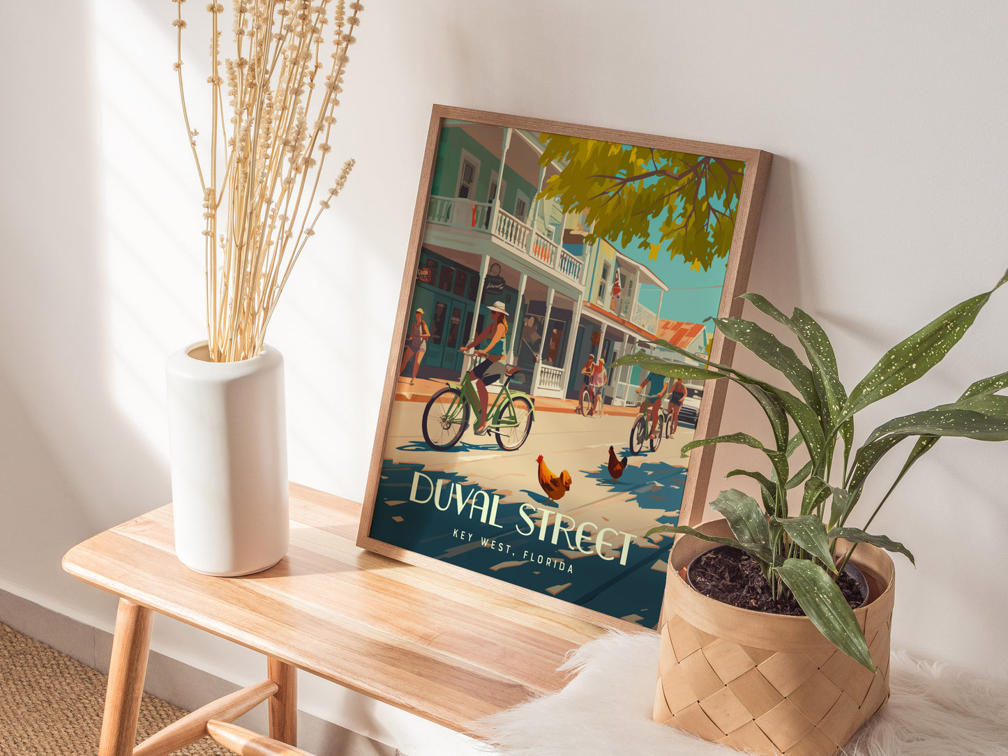 A colorful poster of Duval Street, Key West, featuring cyclists and roosters, rests on a wooden bench beside a plant and a vase with dried flowers.