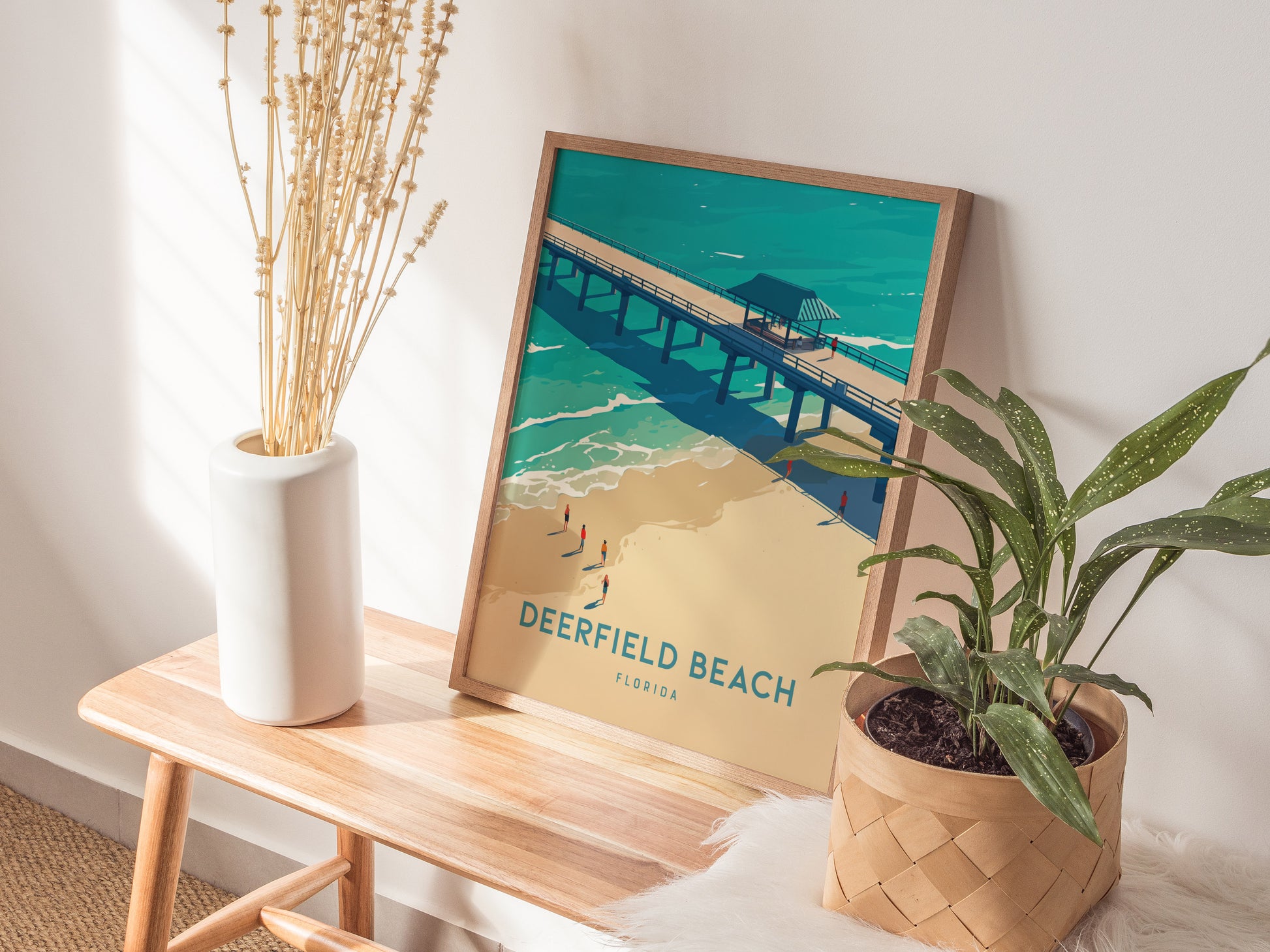 A framed Deerfield Beach poster rests on a wooden bench next to a vase with dried flowers and a potted plant, creating a serene.