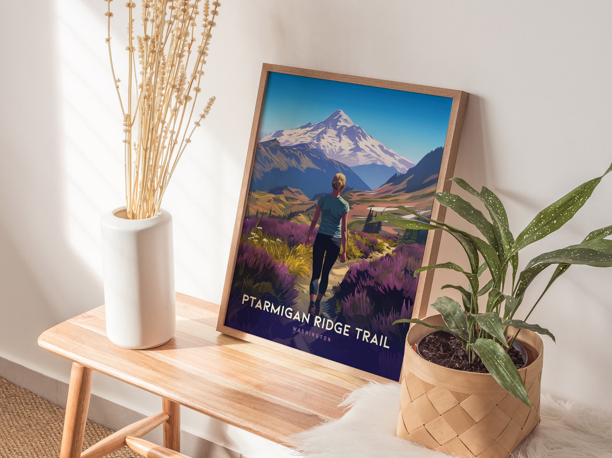 A framed Ptarmigan Ridge Trail poster leans against a wall, surrounded by a white vase with dried flowers and a potted plant on a wooden bench.