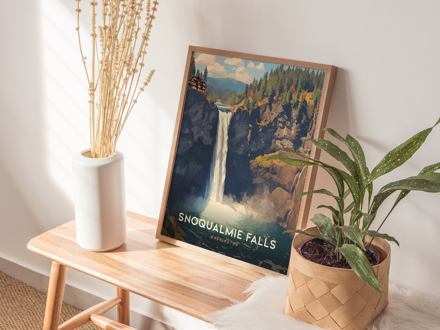 A framed Snoqualmie Falls poster rests on a wooden bench beside a white vase with dried flowers and a potted green plant, creating a cozy, natural decor setup.