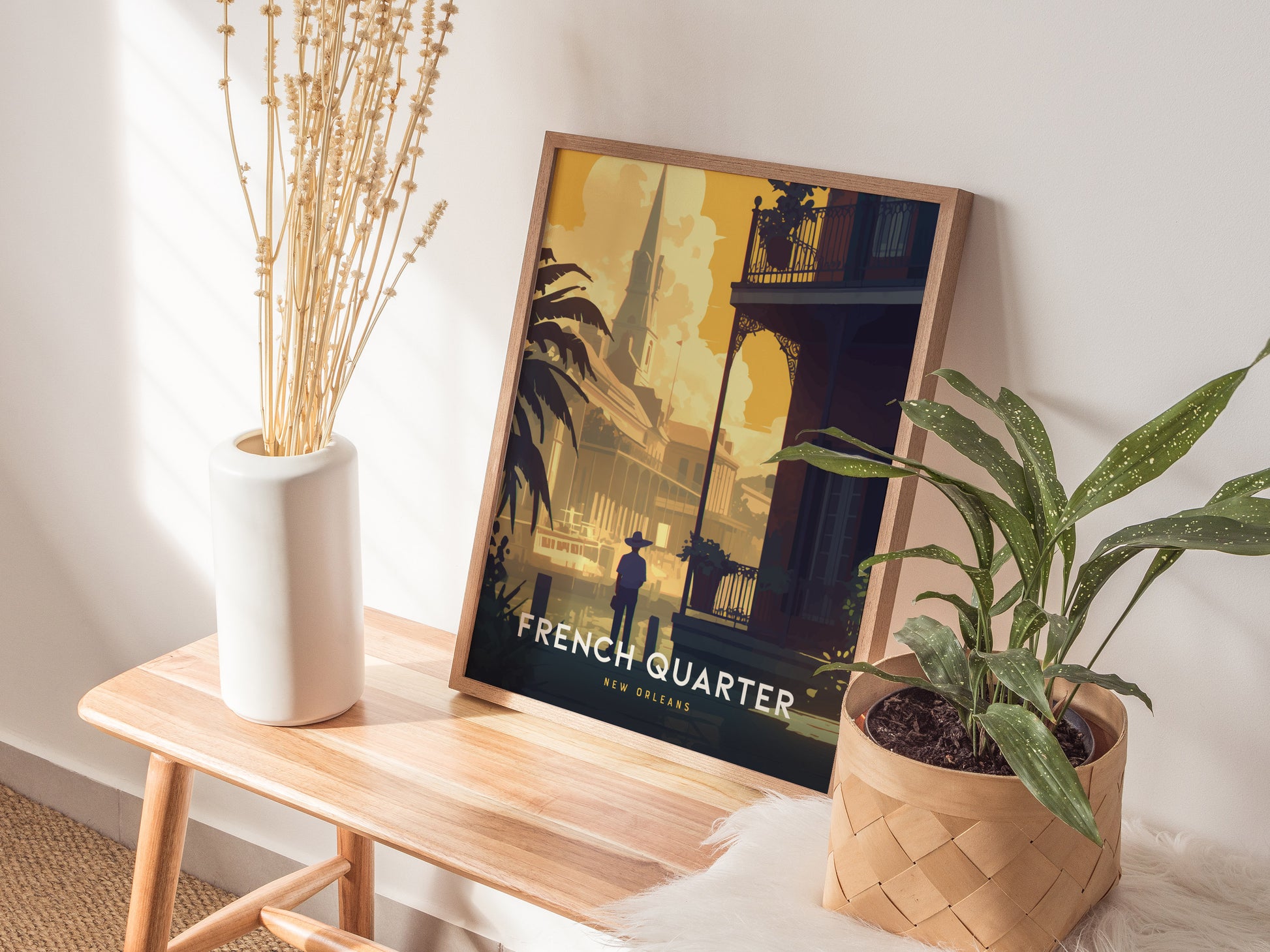 A framed poster of the French Quarter, New Orleans, rests on a wooden bench beside a white vase with dried flowers and a potted plant.