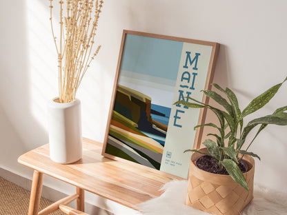 A framed minimalist poster with Maine text rests on a wooden bench, accompanied by a white vase with dried flowers and a potted plant. Sunlight casts soft shadows.