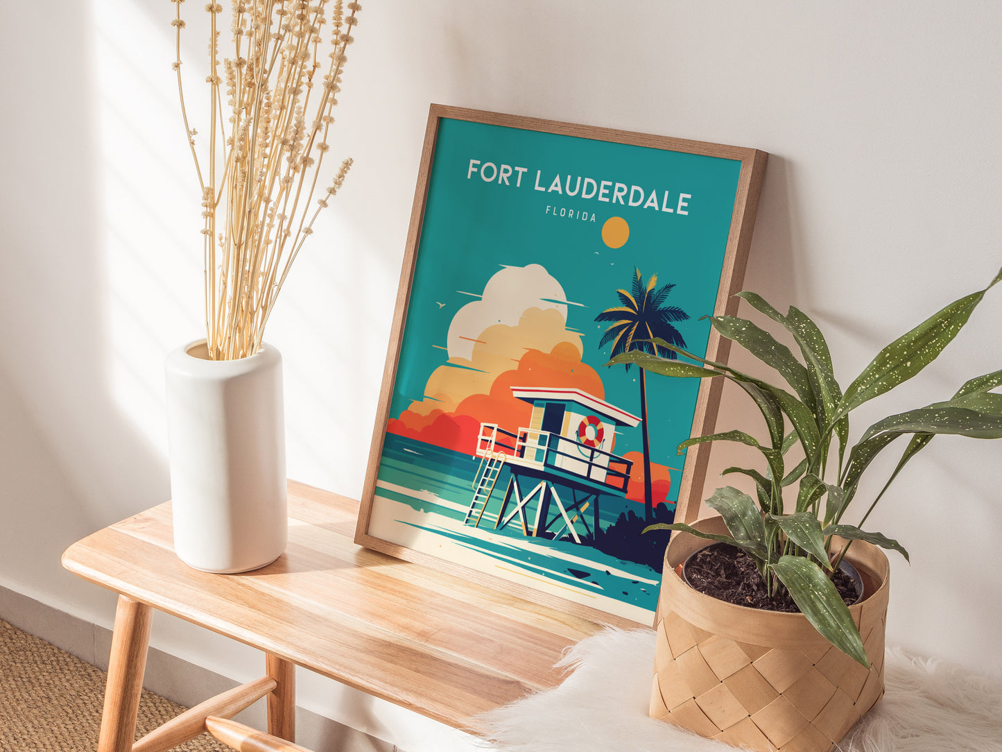 A framed Fort Lauderdale poster featuring a colorful beach scene with a lifeguard stand and palm tree, placed on a wooden bench next to a potted plant and vase.