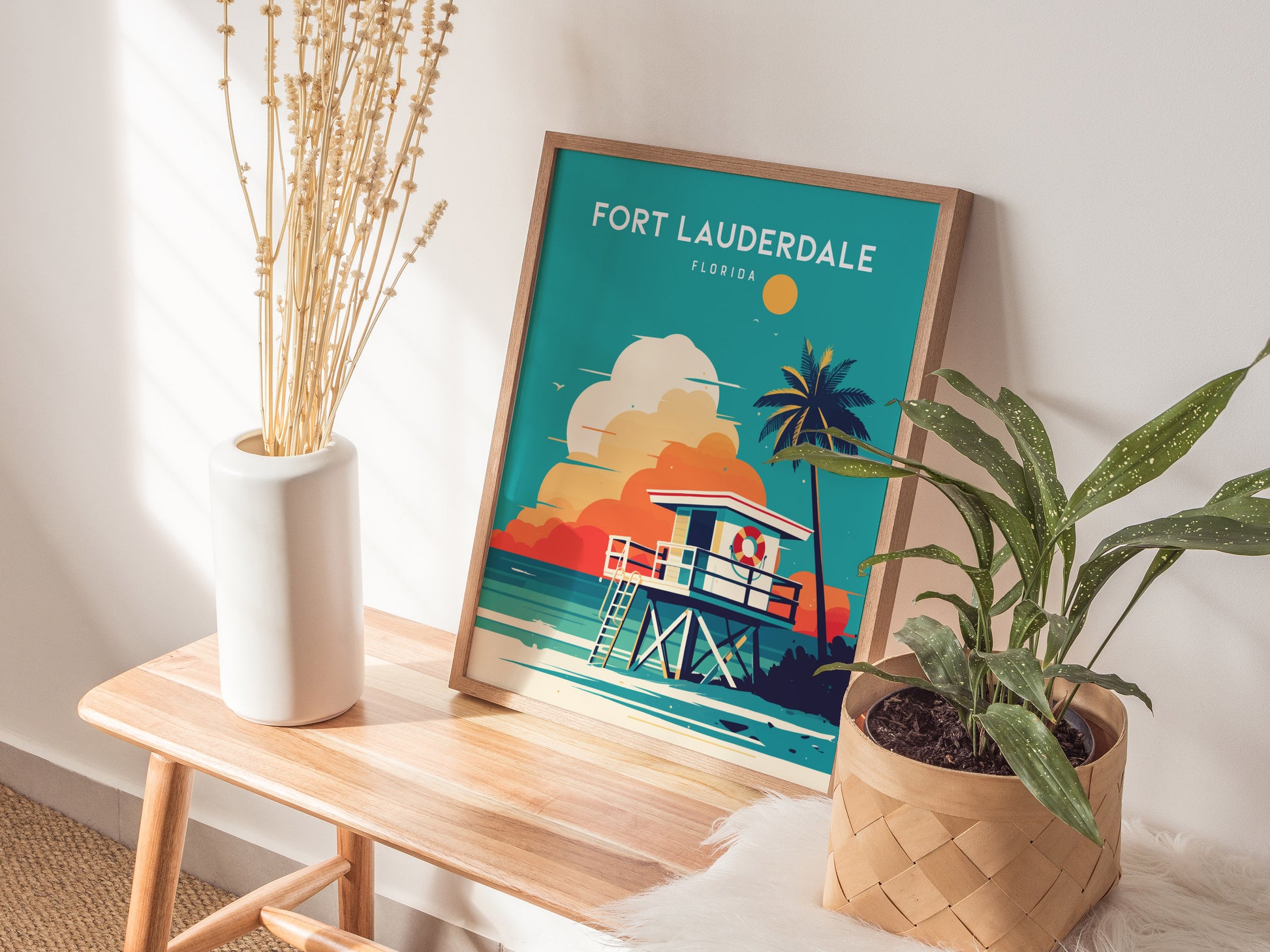A framed Fort Lauderdale poster featuring a colorful beach scene with a lifeguard stand and palm tree, placed on a wooden bench next to a potted plant and vase.