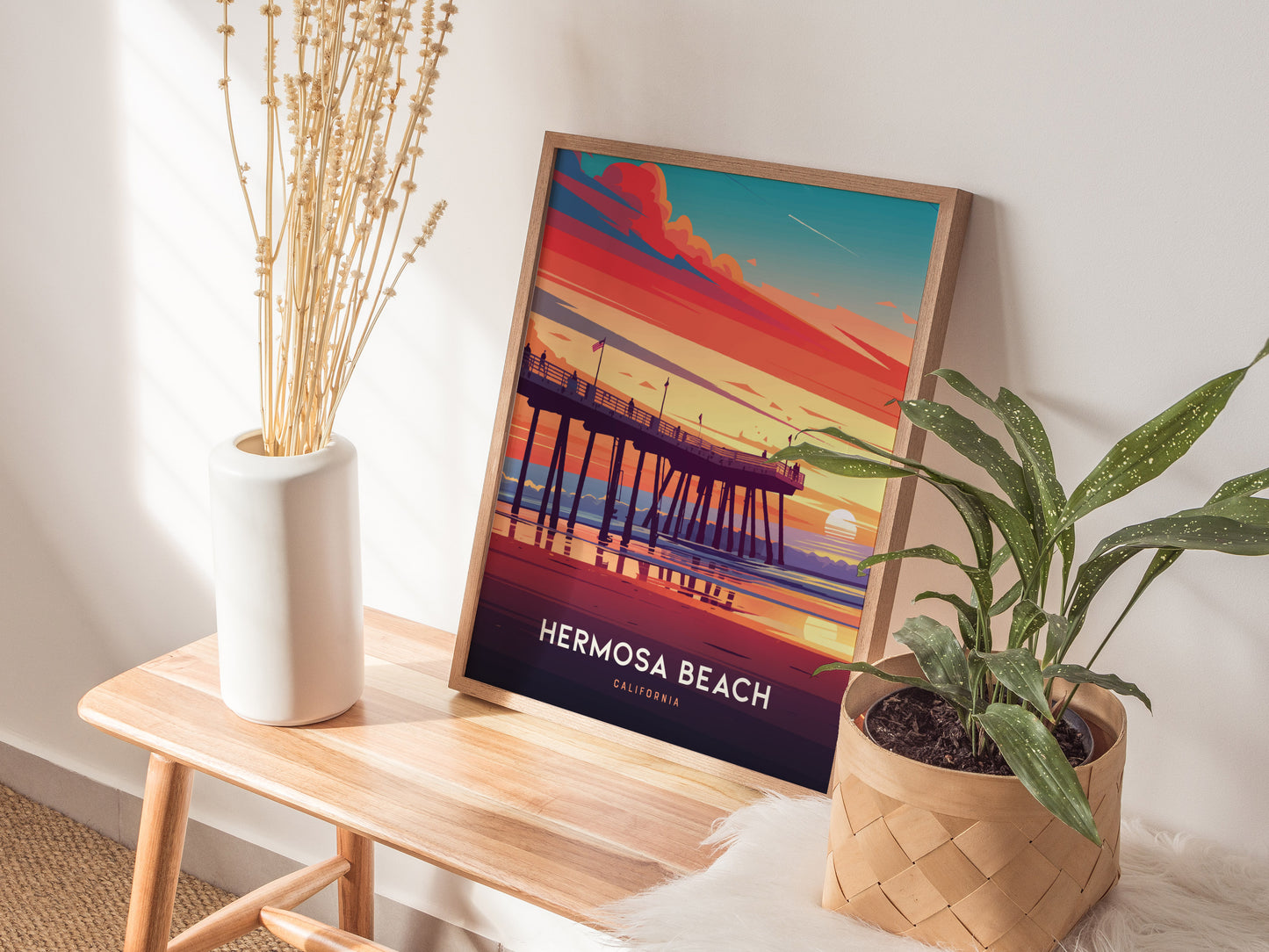 A framed Hermosa Beach poster depicting a colorful sunset over a pier is displayed on a wooden bench, accompanied by a white vase with dried flowers and a potted plant.