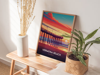 A framed Hermosa Beach poster depicting a colorful sunset over a pier is displayed on a wooden bench, accompanied by a white vase with dried flowers and a potted plant.