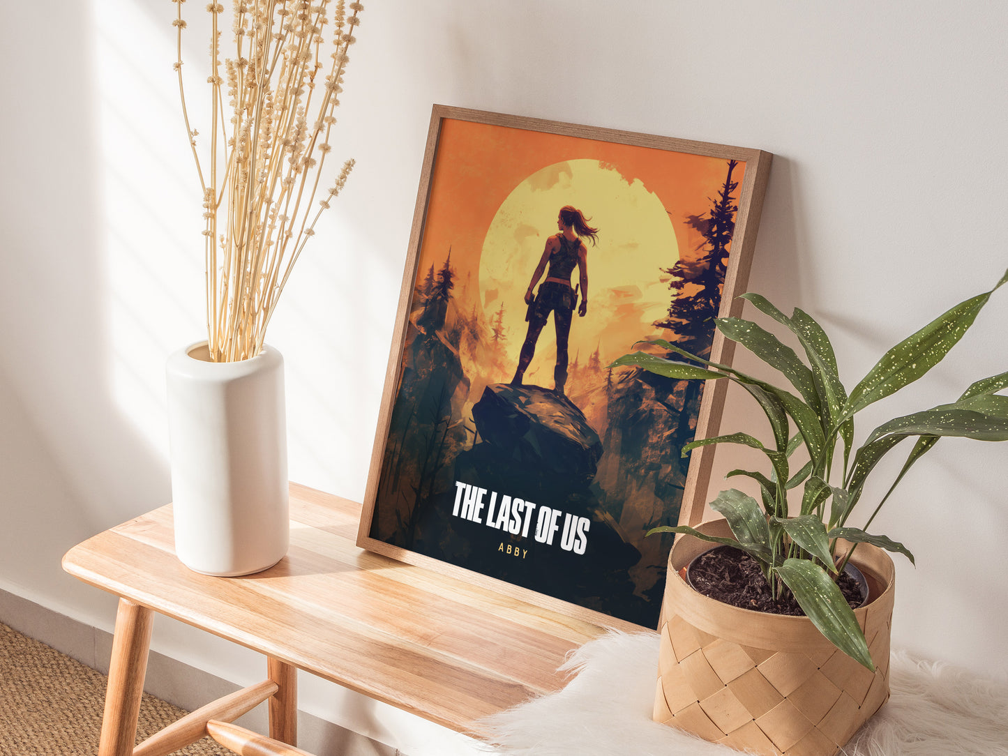 A framed poster of The Last of Us Part 2 featuring Abby stands against a wall, surrounded by a white vase with dried flowers and a potted plant on a.