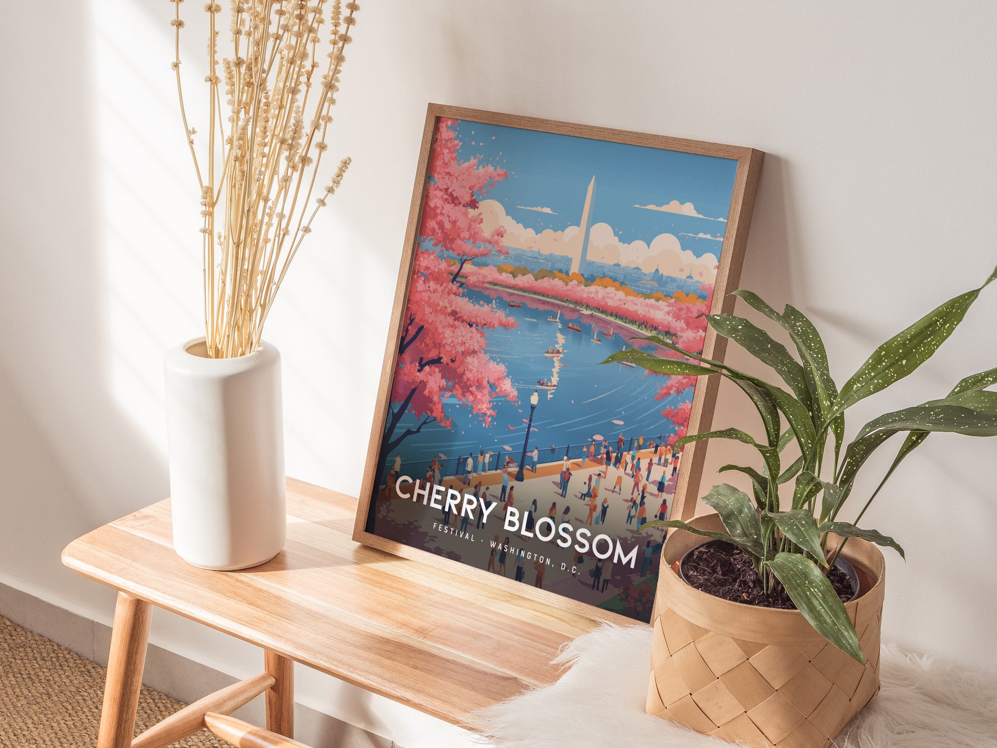 A framed poster depicting the National Cherry Blossom Festival with pink blossoms and the Washington Monument is placed on a wooden bench. It is flanked by a tall white vase with dried stems and a green potted plant.