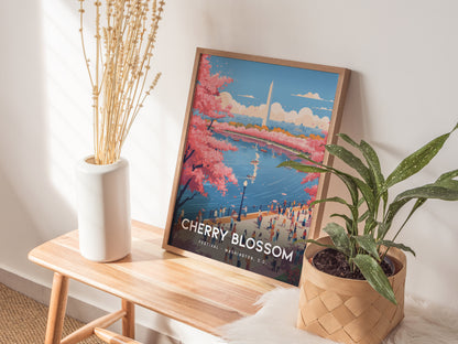 A framed poster depicting the National Cherry Blossom Festival with pink blossoms and the Washington Monument is placed on a wooden bench. It is flanked by a tall white vase with dried stems and a green potted plant.