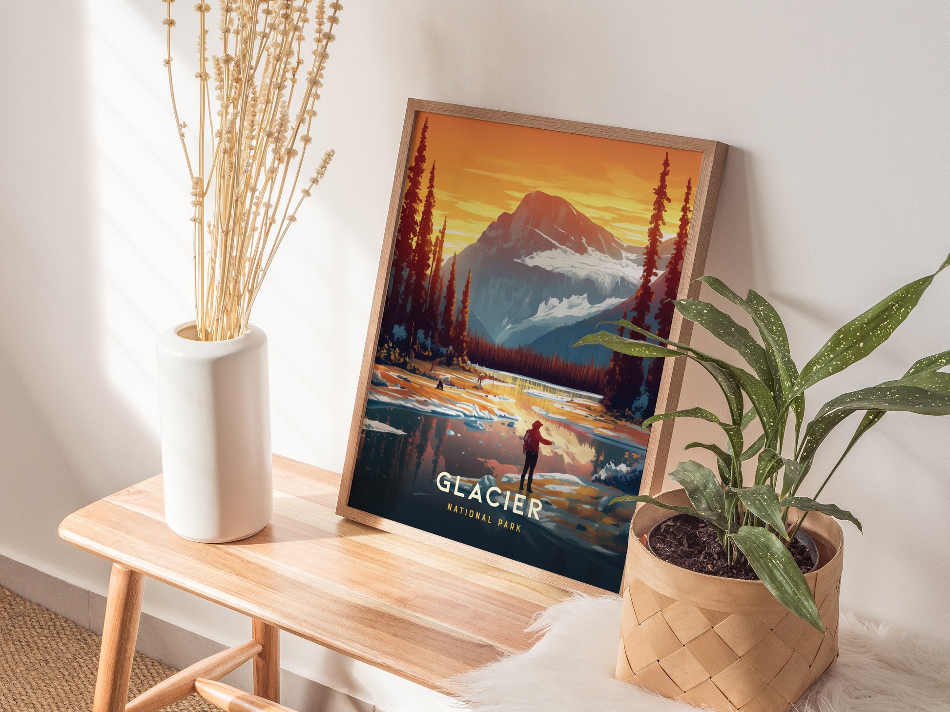 A framed Glacier National Park poster rests on a wooden bench beside a white vase with dried flowers and a potted green plant, creating a cozy, nature-inspired setting.