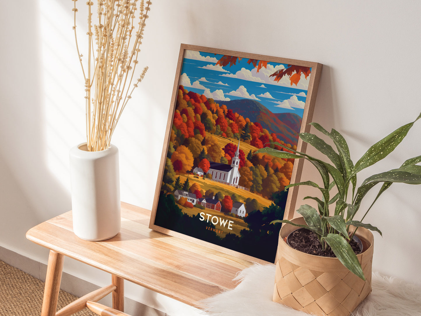 A framed poster of Stowe, Vermont, showcasing a white church steeple surrounded by vibrant fall foliage, rests on a wooden bench beside a vase of dried stems and a potted.