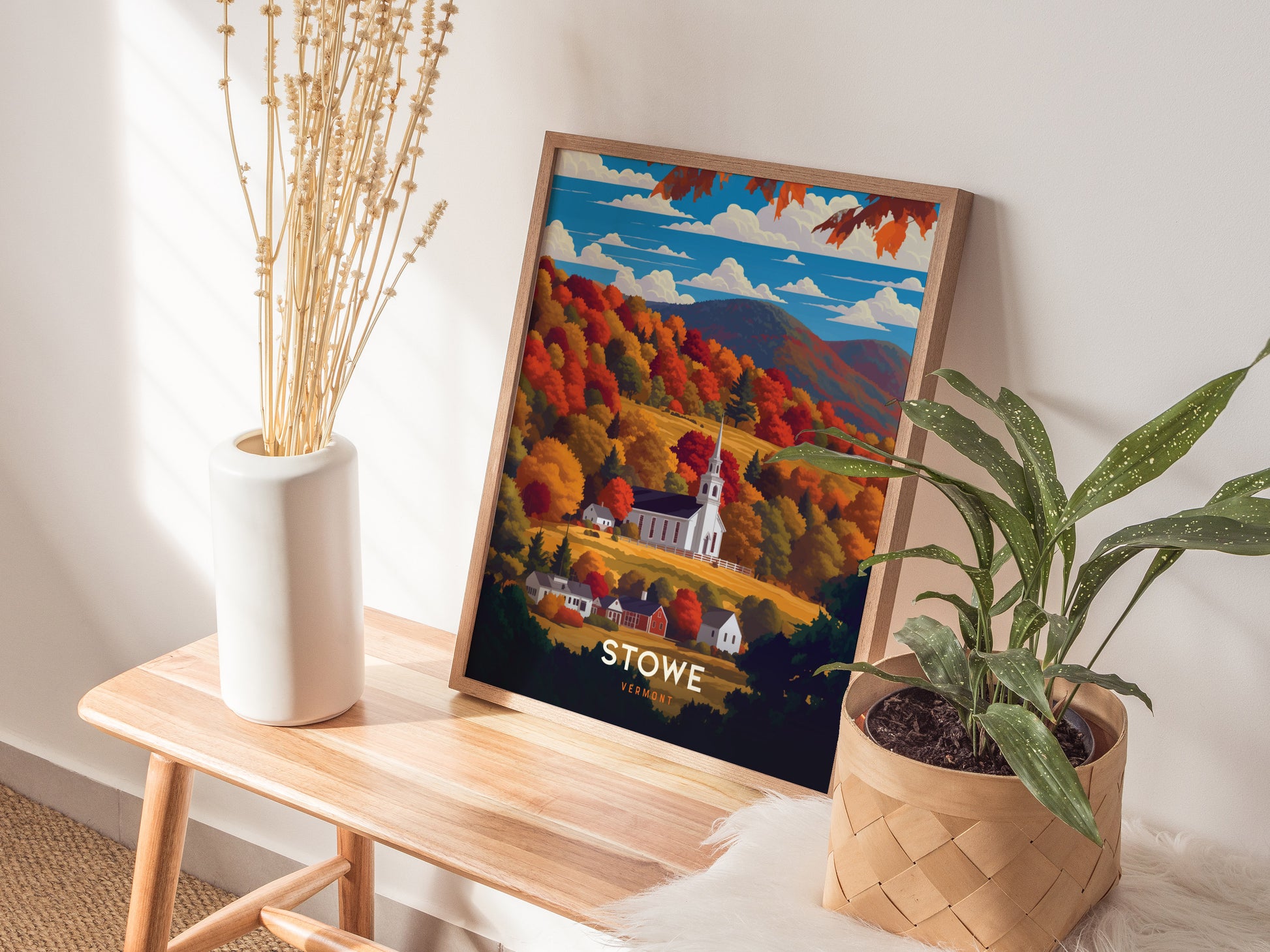 A framed poster of Stowe, Vermont, showcasing a white church steeple surrounded by vibrant fall foliage, rests on a wooden bench beside a vase of dried stems and a potted.
