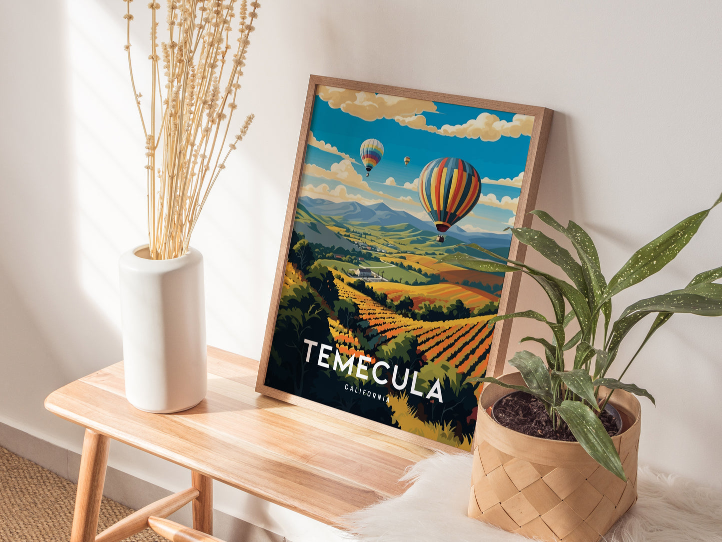 A Temecula poster featuring colorful hot air balloons over vineyards is framed on a wooden bench, accompanied by a white vase with dried stems and a potted plant.