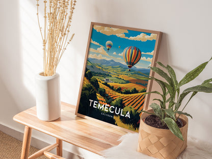 A Temecula poster featuring colorful hot air balloons over vineyards is framed on a wooden bench, accompanied by a white vase with dried stems and a potted plant.