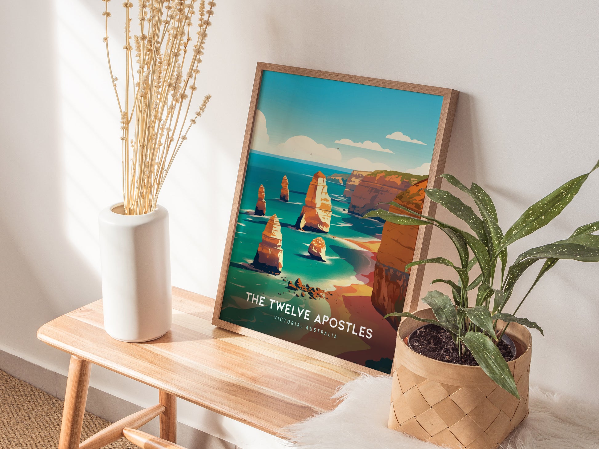 A framed poster of the Twelve Apostles in Australia rests on a wooden bench beside a white vase with dried flowers and a potted green plant.