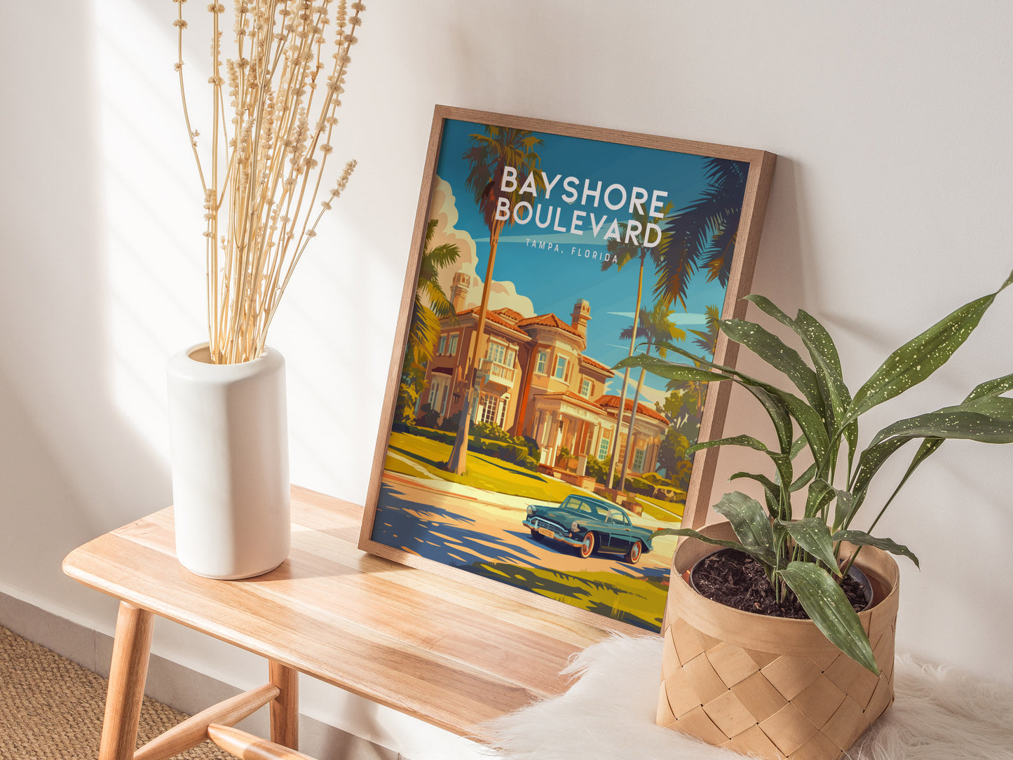 A Bayshore Boulevard Tampa poster rests on a wooden bench, surrounded by a white vase with dried stems and a green potted plant, bathed in sunlight.