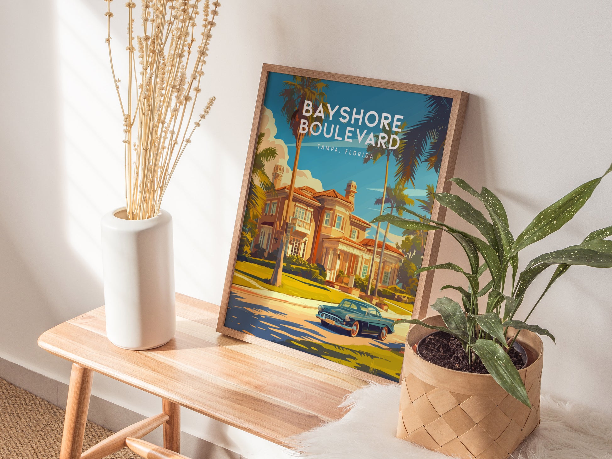 A Bayshore Boulevard Tampa poster rests on a wooden bench, surrounded by a white vase with dried stems and a green potted plant, bathed in sunlight.