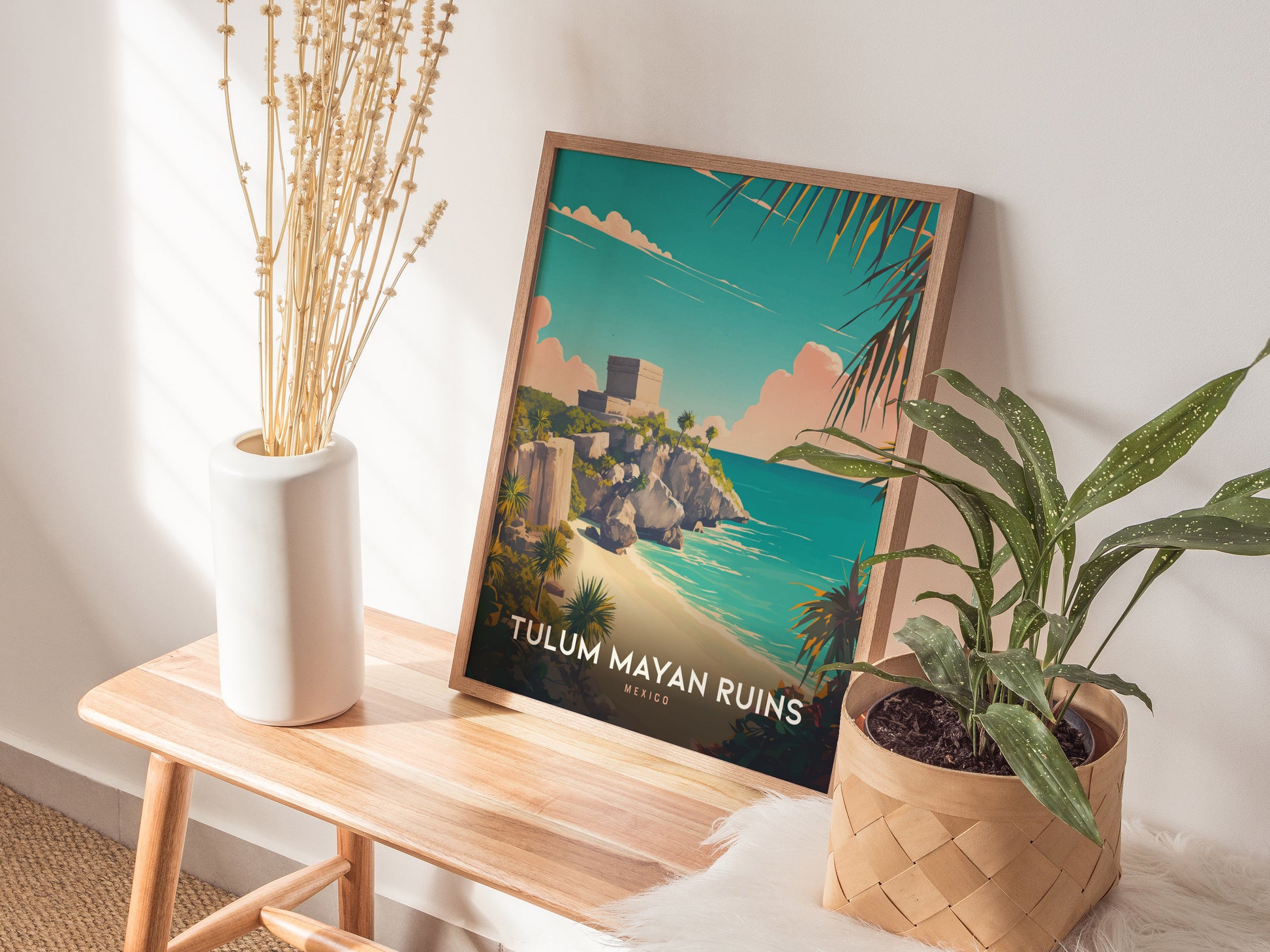 A framed Tulum Mayan Ruins poster rests on a wooden bench, surrounded by a white vase with dried flowers and a potted green plant, against a softly lit wall.
