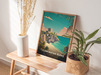 A framed Tulum Mayan Ruins poster rests on a wooden bench, surrounded by a white vase with dried flowers and a potted green plant, against a softly lit wall.