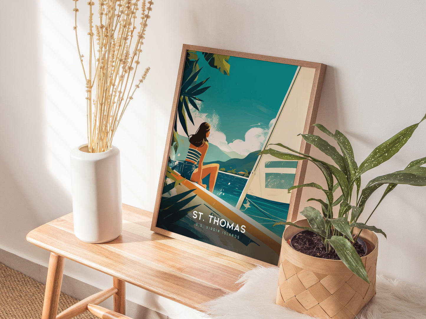 A framed St. Thomas poster in a retro travel style sits on a wooden bench, flanked by a white vase with dried flowers and a potted plant.