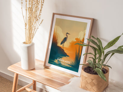 A framed Everglades poster featuring a heron by water is displayed on a wooden bench, accompanied by a white vase with dried flowers and a potted plant.