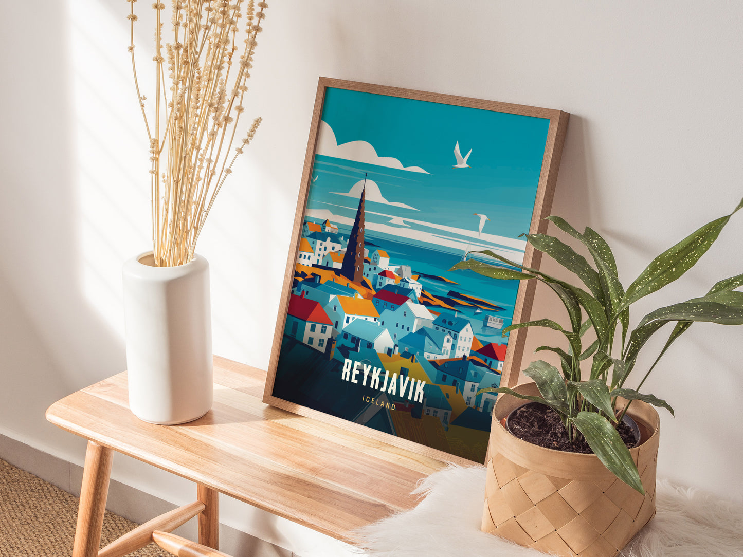 A framed poster of Reykjavík with colorful buildings and a church is displayed on a wooden bench, accompanied by a white vase with dried stems and a potted green plant.