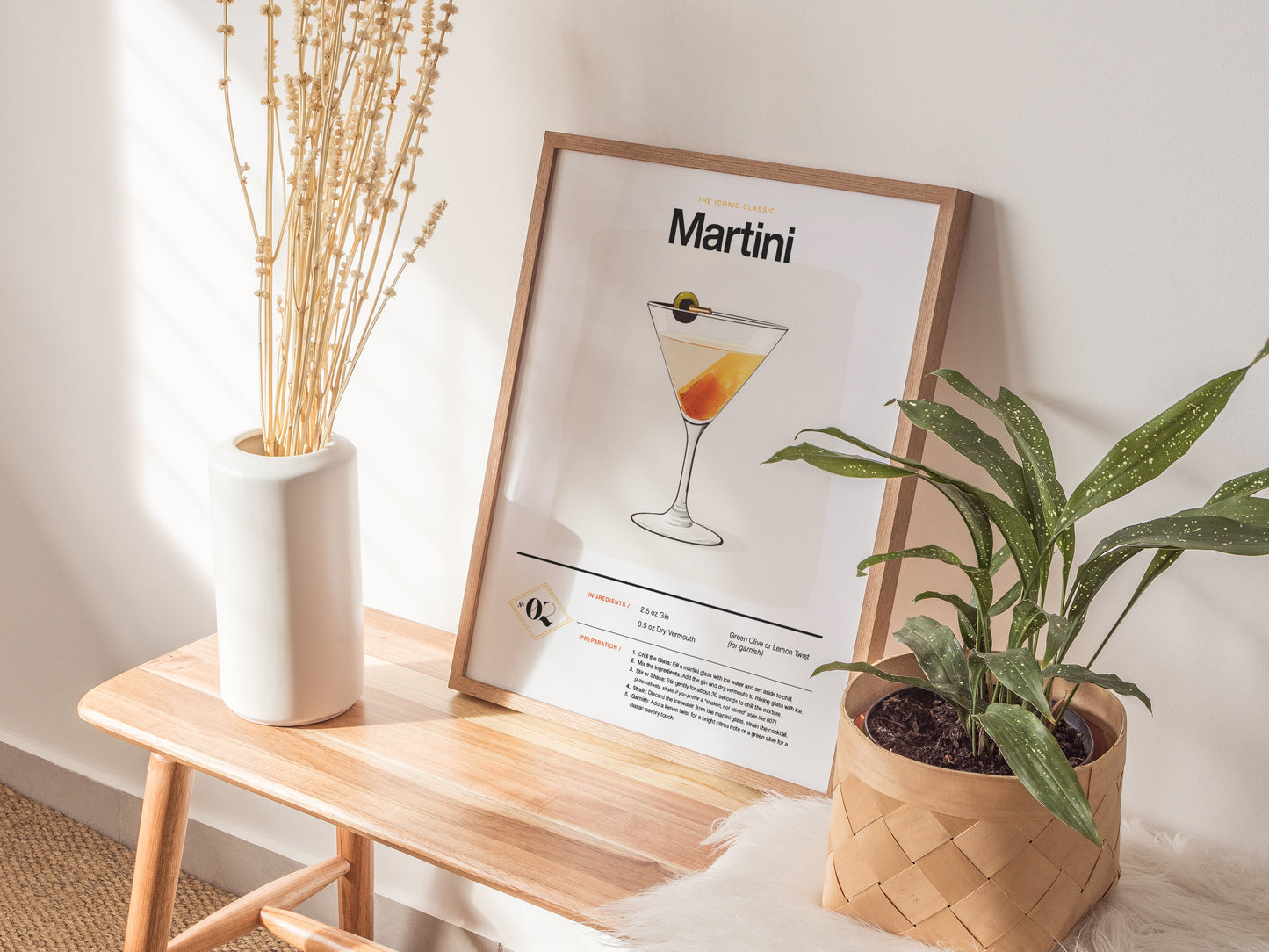 A framed Martini cocktail poster rests on a wooden bench, accompanied by a white vase with dried stems and a potted green plant, creating a stylish interior decor setup.
