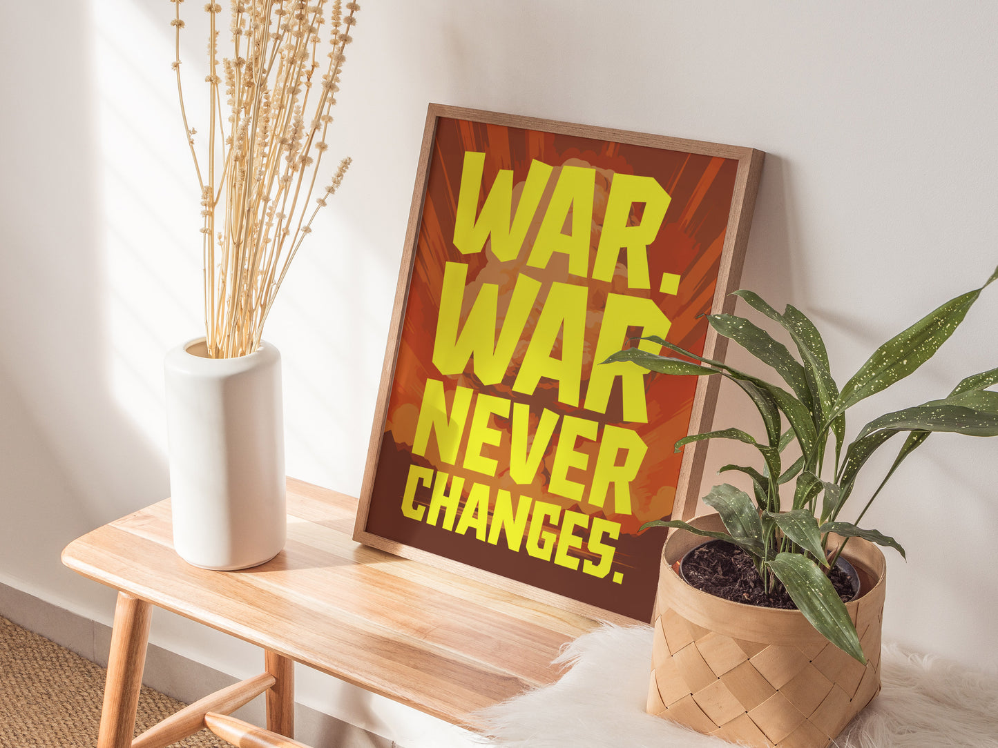A framed poster with bold yellow text WAR. WAR NEVER CHANGES. rests on a wooden bench next to a white vase with dried stems and a green potted plant.