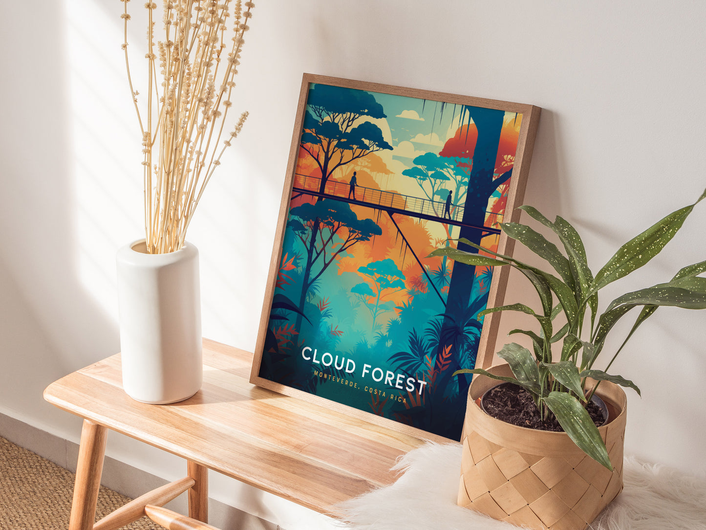 A colorful poster of Costa Ricas Cloud Forest, featuring a canopy walkway, is propped on a wooden bench beside a white vase with dried stems and a leafy plant.