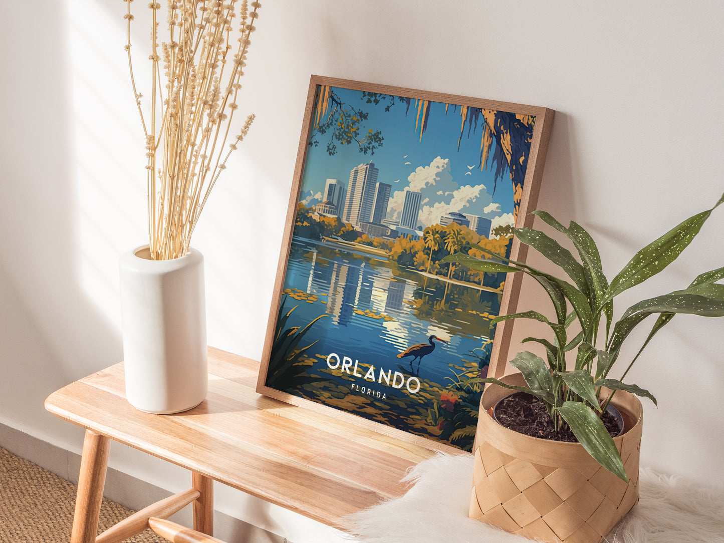 A framed poster depicting Orlandos skyline with Lake Eola is propped on a wooden bench, surrounded by a white vase of dried flowers and a potted plant.