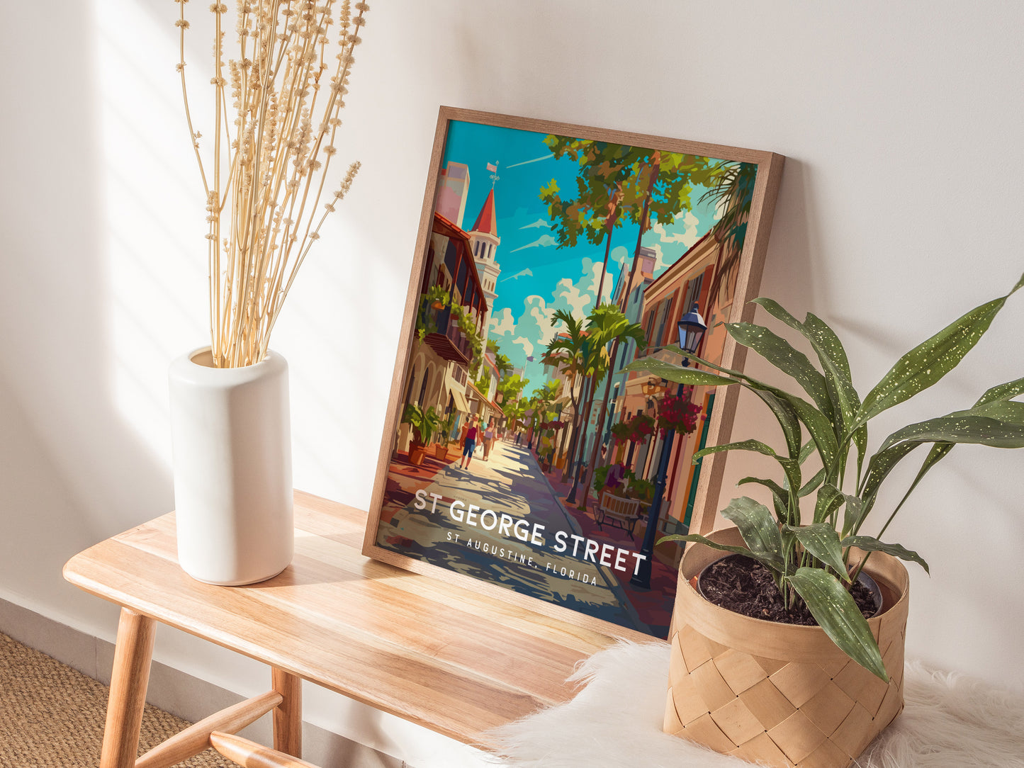 A framed St. George Street poster leans against a wall, surrounded by a white vase with dried branches and a potted plant on a wooden bench.