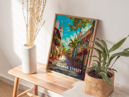 A framed St. George Street poster leans against a wall, surrounded by a white vase with dried branches and a potted plant on a wooden bench.