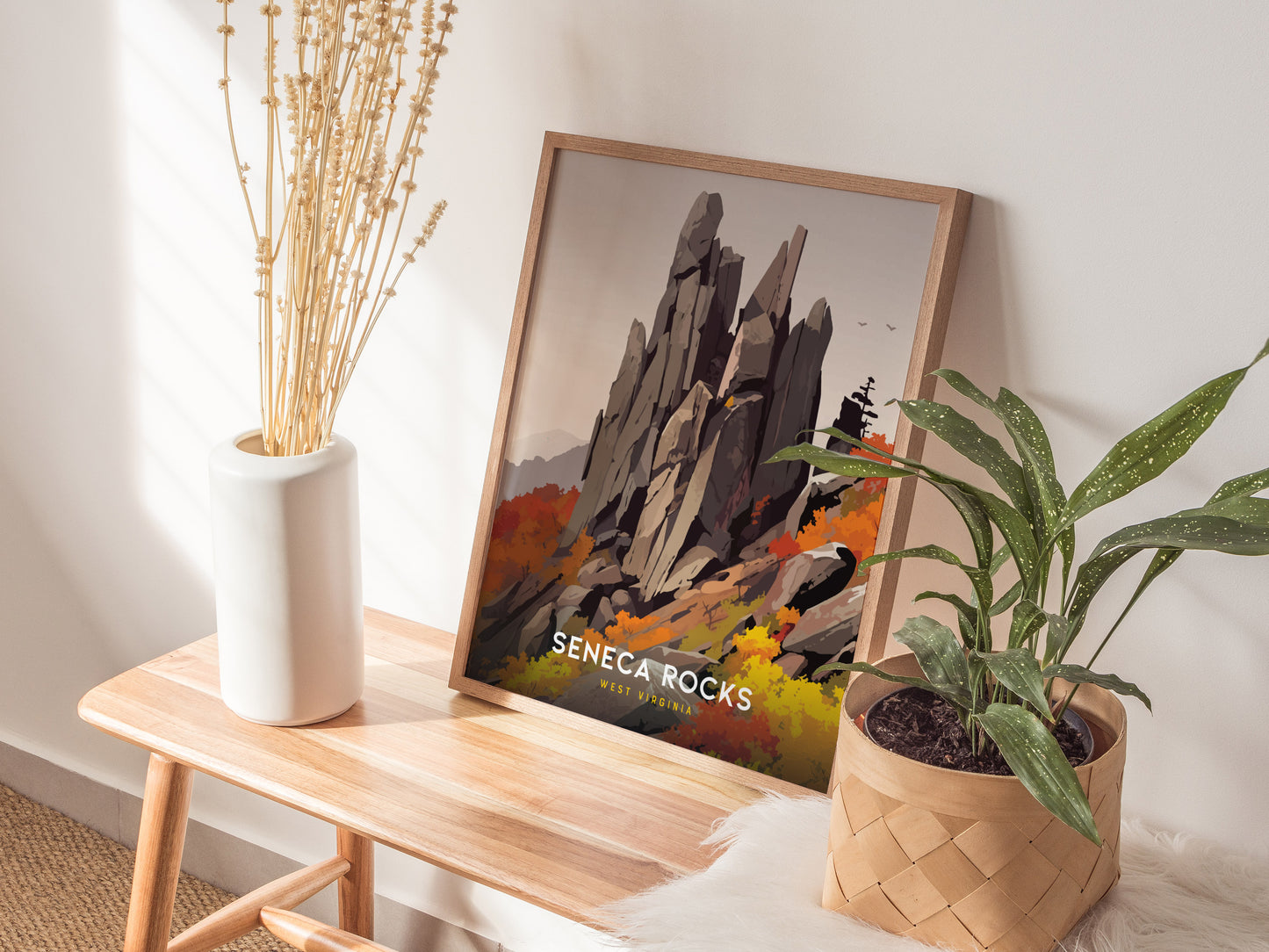 A framed Seneca Rocks poster sits on a wooden bench, surrounded by a white vase with dried flowers and a potted green plant. Sunlight casts shadows on the wall.