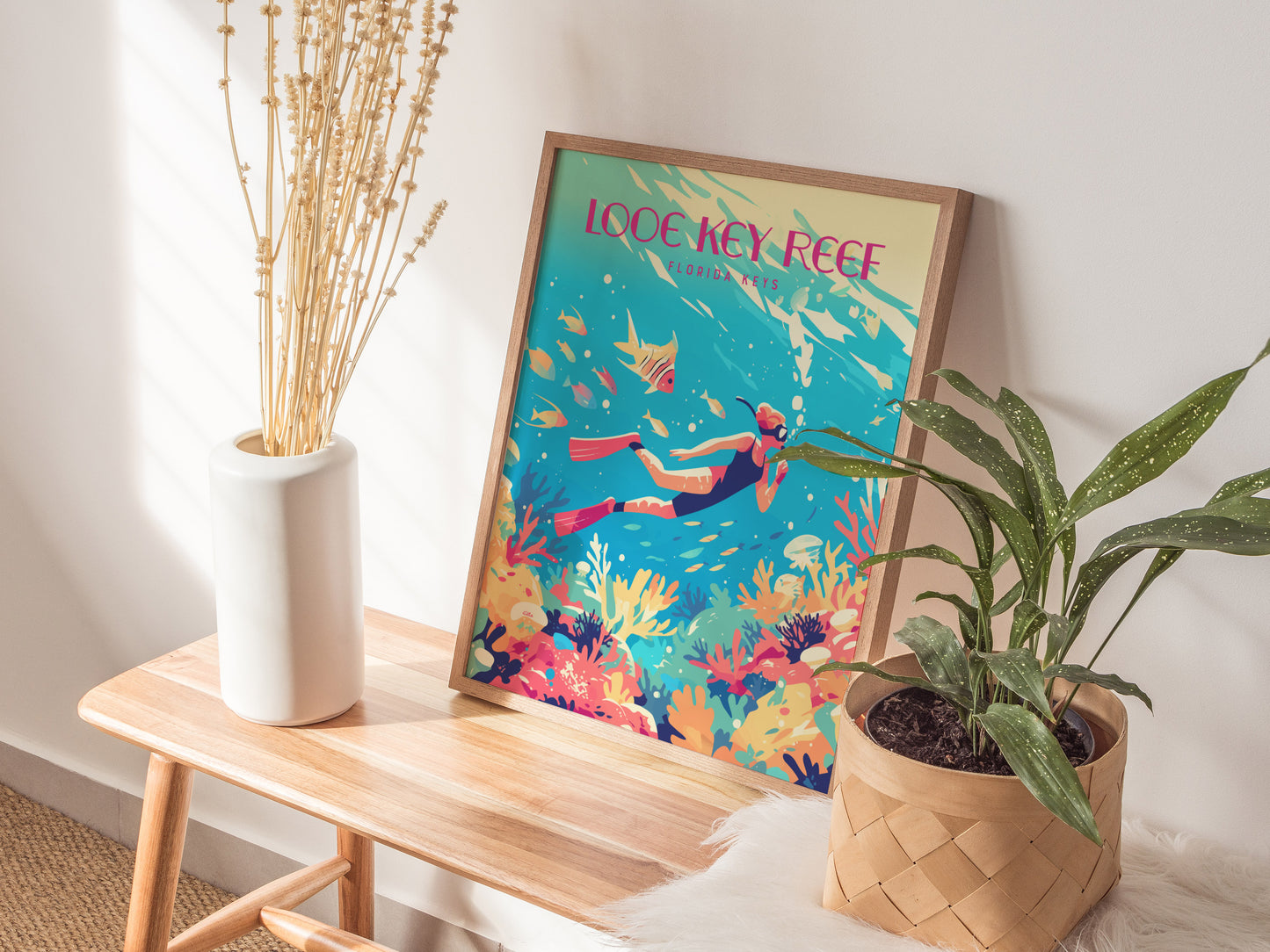 A colorful Looe Key Reef poster with vibrant coral and fish is displayed on a wooden bench, alongside a white vase with dried flowers and a potted green plant.