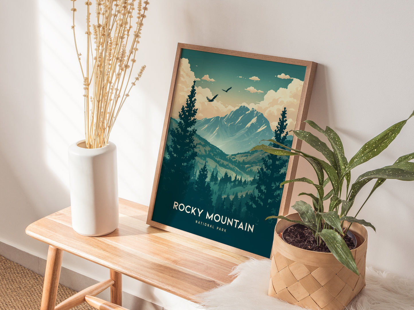 A framed Rocky Mountain National Park poster rests on a wooden bench, surrounded by a white vase with dried flowers and a potted green plant.