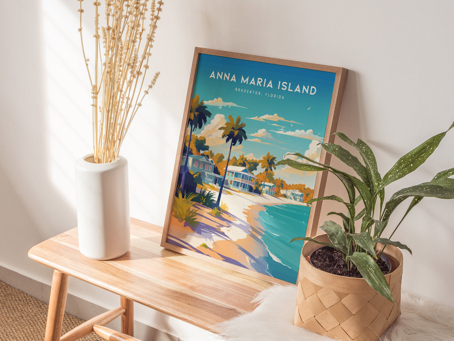 A wooden bench holds a framed Anna Maria Island poster depicting a colorful beach scene with palm trees. Beside it, a white vase with tall dried stems and a woven basket with a green plant add a natural touch.