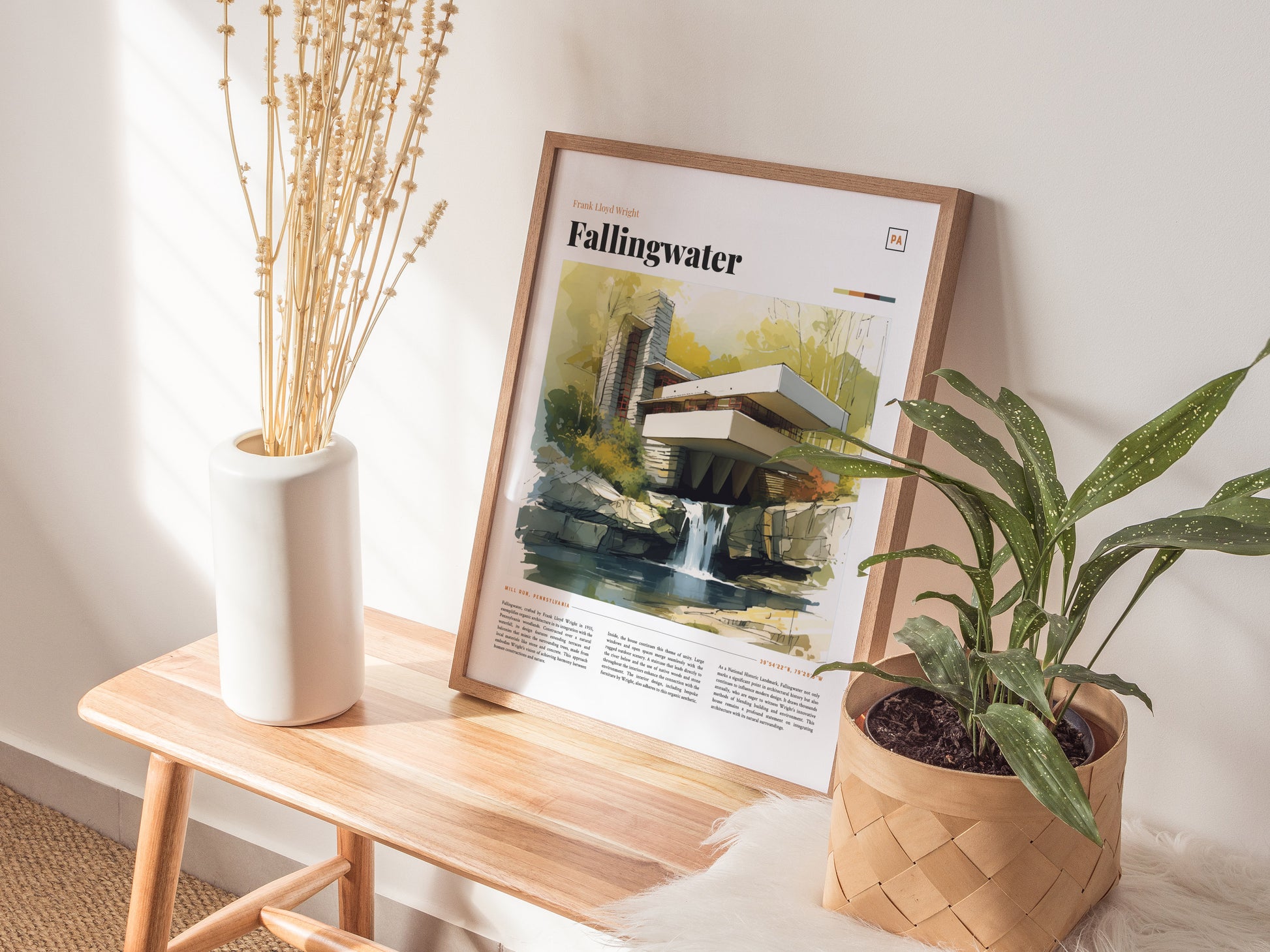 A framed Fallingwater poster rests on a wooden bench, accompanied by a white vase with dried stems and a potted green plant.