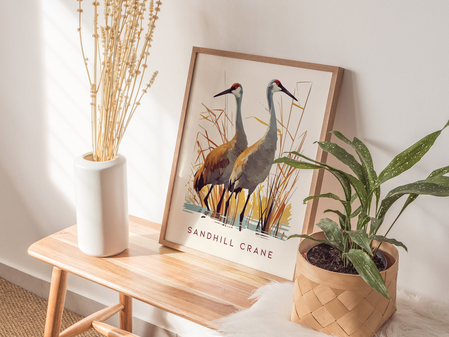 A framed Sandhill Crane poster rests on a wooden bench, accompanied by a white vase with dried flowers and a potted green plant, creating a serene decor setting.