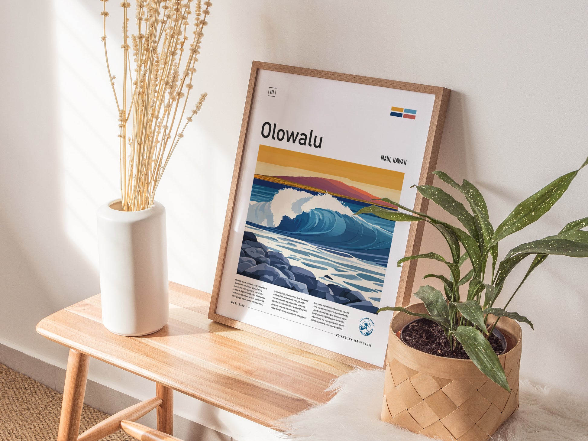 A framed Olowalu Maui surf poster leans against a wall on a wooden bench, accompanied by a white vase with dried flowers and a potted plant.
