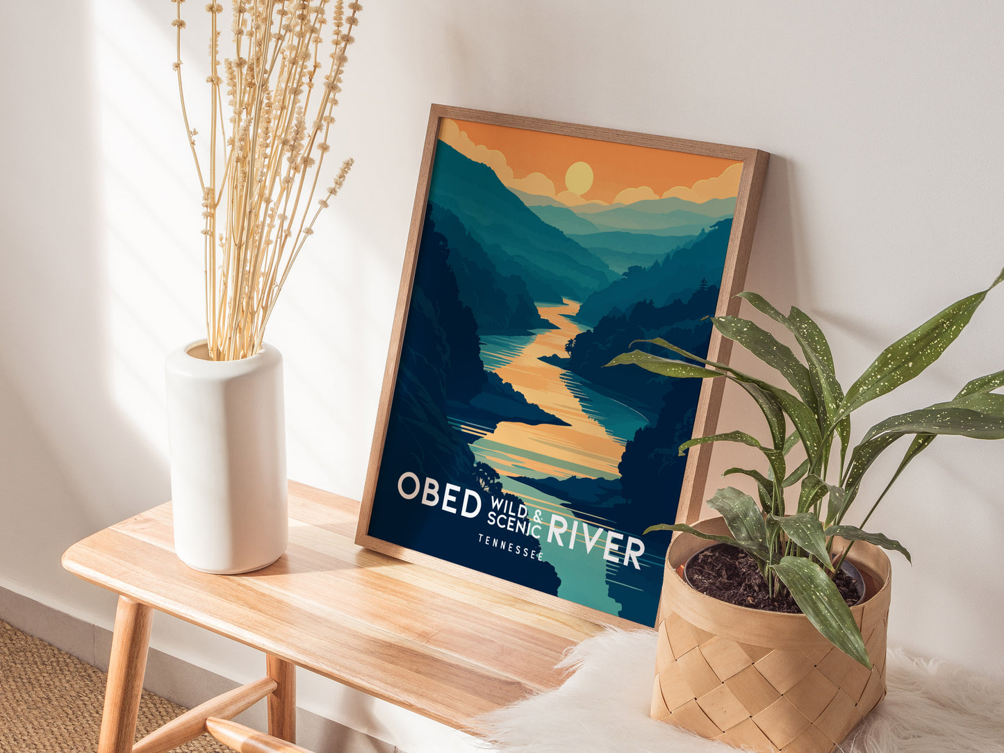 A framed Obed Wild and Scenic River poster sits on a wooden bench, accompanied by a white vase with dried flowers and a potted plant. Sunlight streams in, creating shadows.