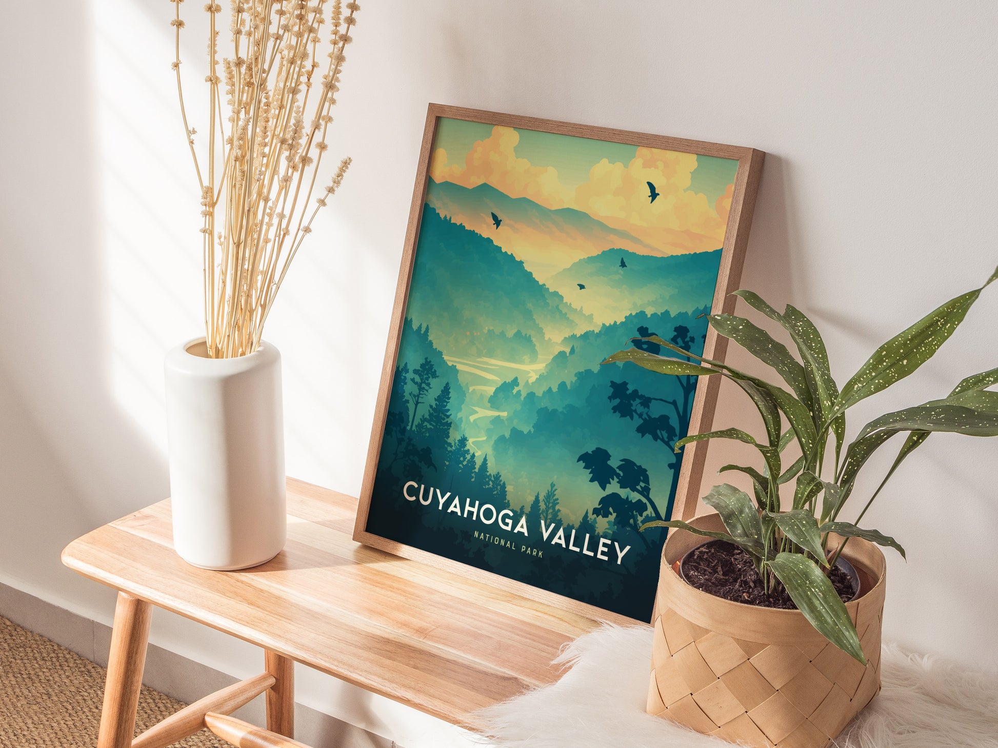 A framed Cuyahoga Valley National Park poster sits on a wooden bench beside a white vase with dried flowers and a potted plant in a woven basket.